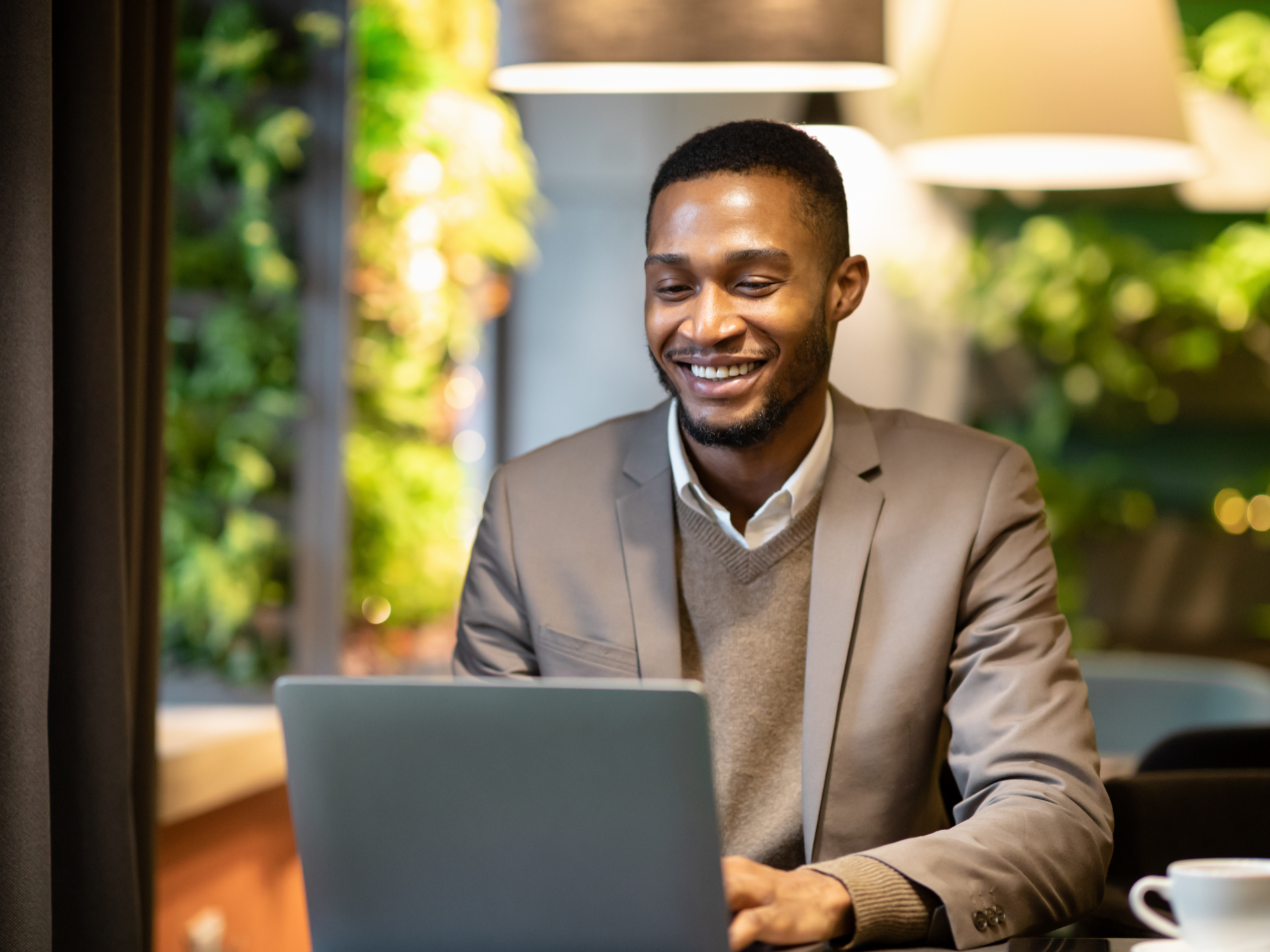 Smiling businessman in a blazer working on a laptop at a modern, plant-filled cafe.